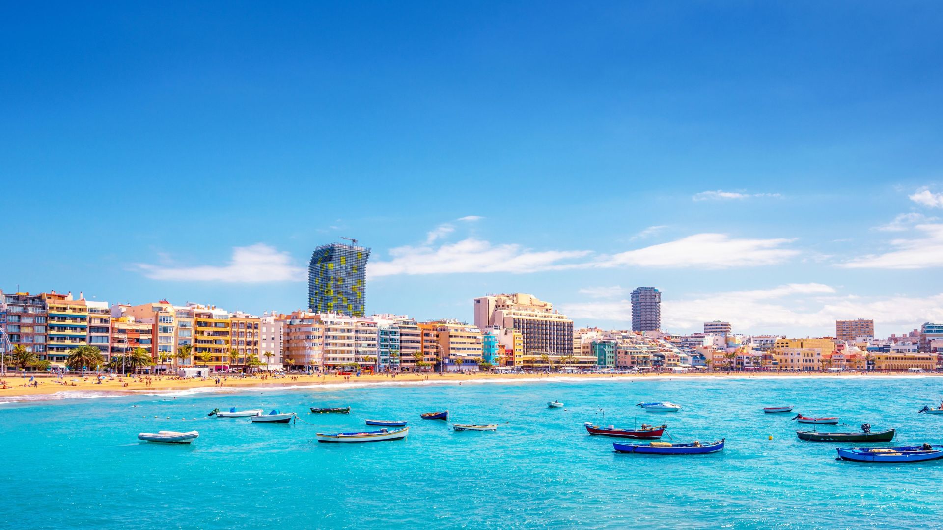 Zicht op een heldere, turquoise zee met kleine boten en een stadsgezicht op de achtergrond onder een blauwe hemel.